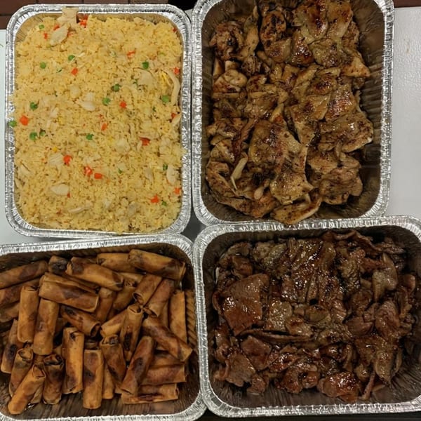 Four foil trays of Vietnamese food, including fried rice, spring rolls, grilled chicken, and beef.