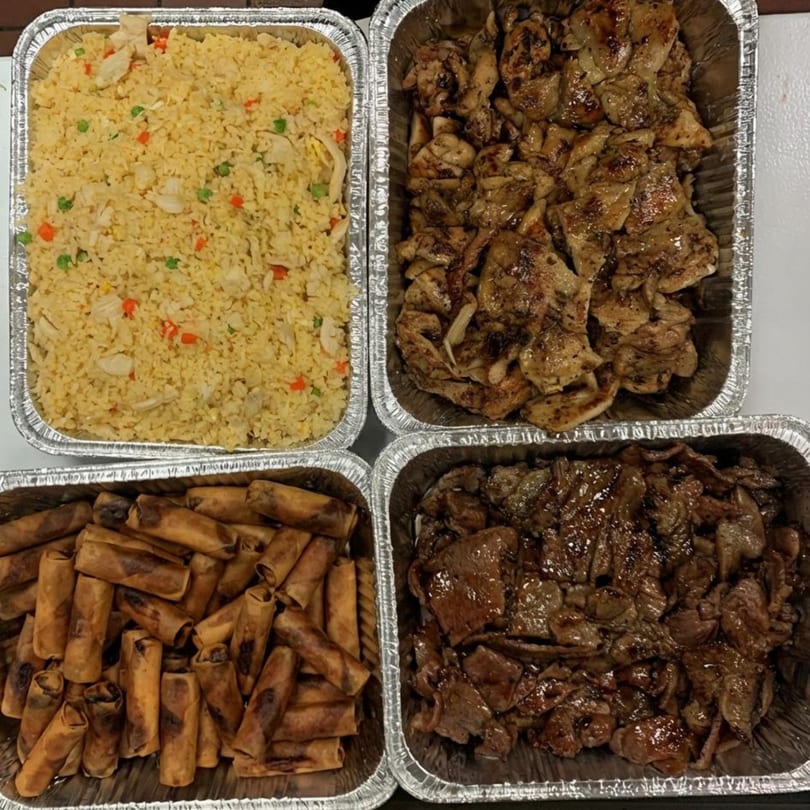 Four foil trays of Vietnamese food, including fried rice, spring rolls, grilled chicken, and beef.